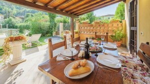 Uma área de refeições ao ar livre com mesa de madeira posta para dois, rodeada por cadeiras, com vista para um jardim e uma parede de tijolos.