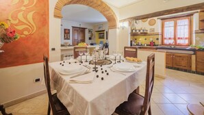 Uma área de refeições com uma mesa posta para quatro pessoas, cozinha ao fundo e parede decorativa com uma grande letra 'S'.