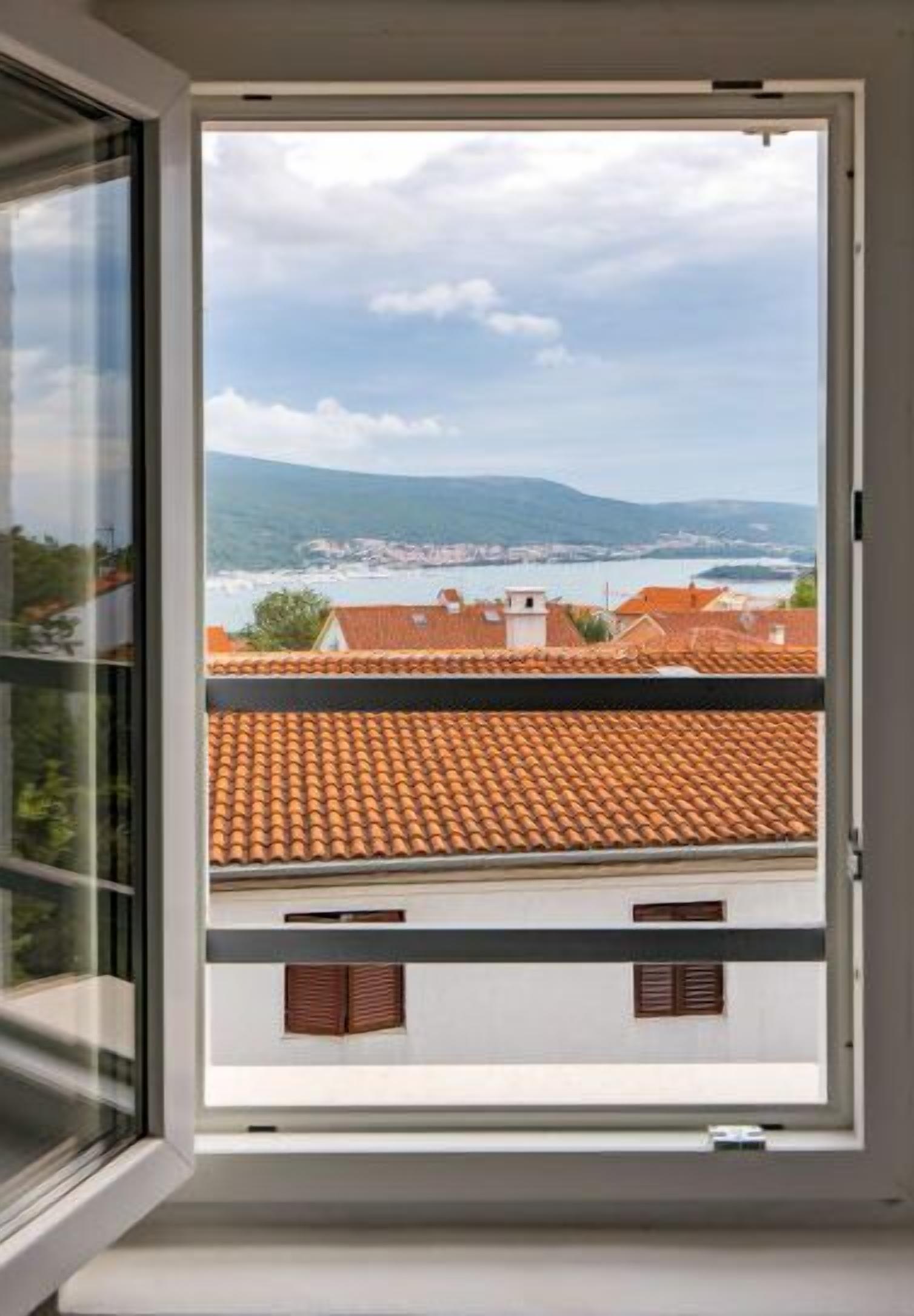 Ein Fenster mit Blick auf eine Küstenstadt, Häuser mit roten Ziegeldächern und ein Gewässer in der Ferne.