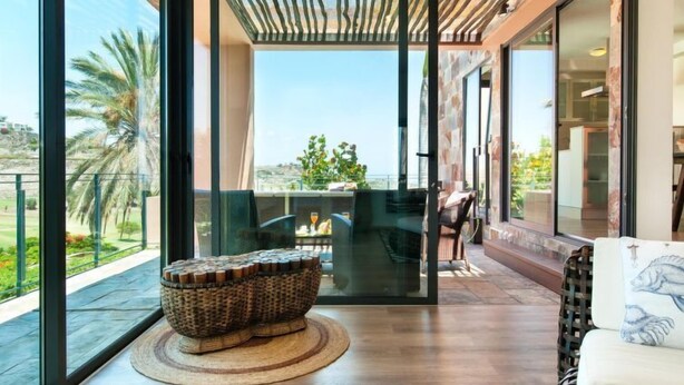 Una sala de estar moderna con un gran ventanal que da a un balcón, una mesa de centro tejida y un sofá con un cojín decorativo.