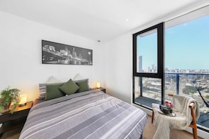 A bedroom with a large bed, a cityscape view, a framed picture on the wall, and a chair by the window. - A Stylish 2BR Apt with City Views in Southbank (Southbank)