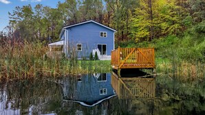 A blue house with a wooden dock extending into a pond, surrounded by trees and vegetation.