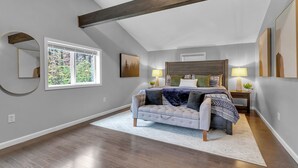 A bedroom with a bed, a sofa, a window with a view of trees, and a ceiling with exposed beams.