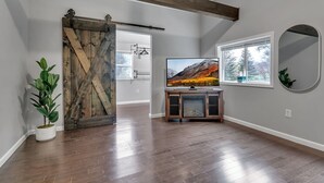 A spacious room with a wooden floor, a sliding barn door, a television on a wooden stand, and a large window with a view of trees.