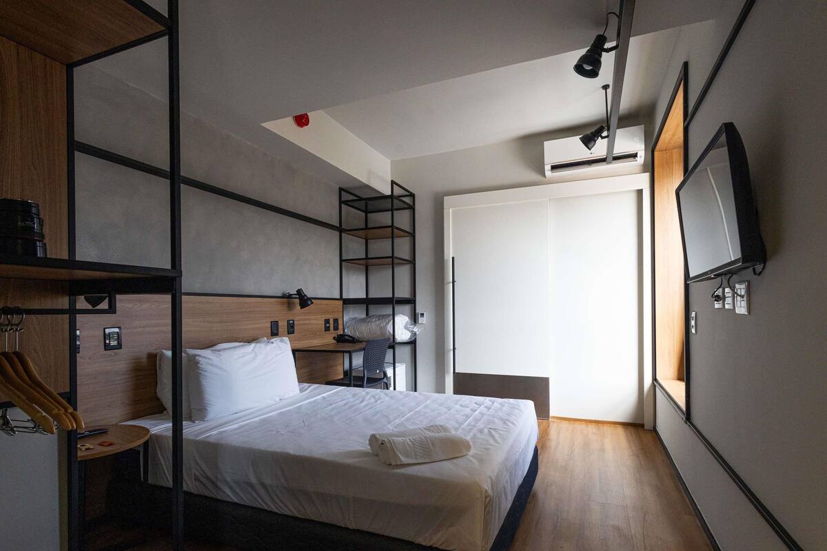 A modern hotel room with a bed, a desk, a chair, a TV, and a wardrobe.
