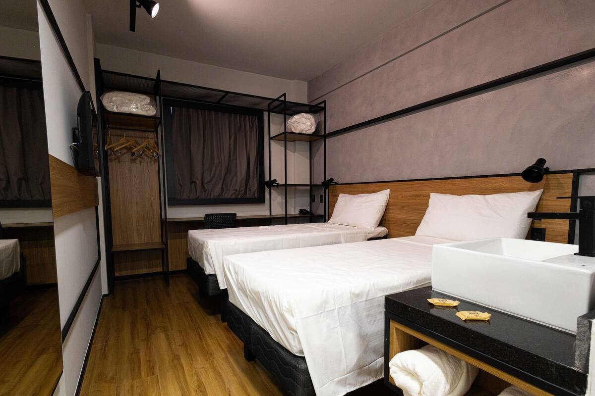 A modern hotel room with two beds, a wooden floor, a built-in desk with a sink, and a large mirror.