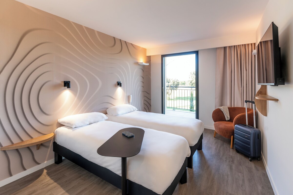 A modern hotel room with two beds, a small table, a chair, a TV, and a window with curtains.