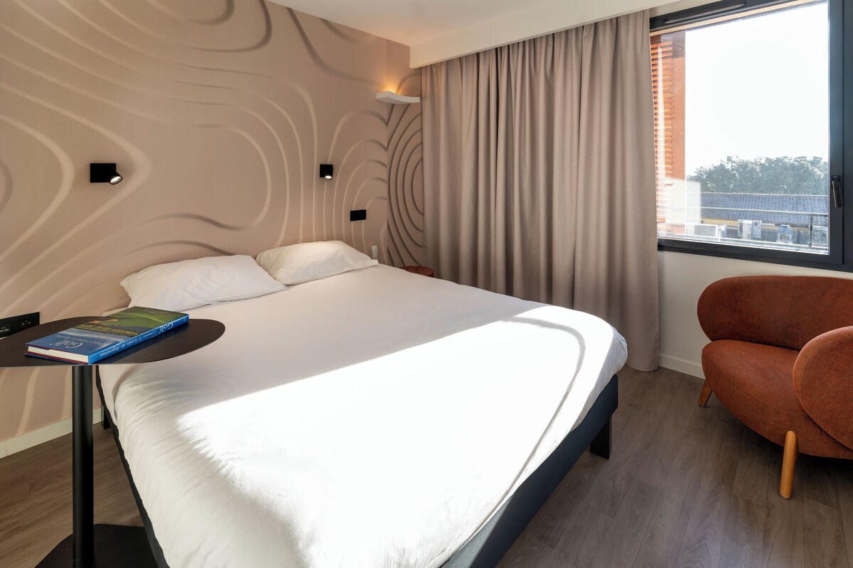 A modern hotel room with a large bed, a small table with a book, a chair, and a window with curtains.
