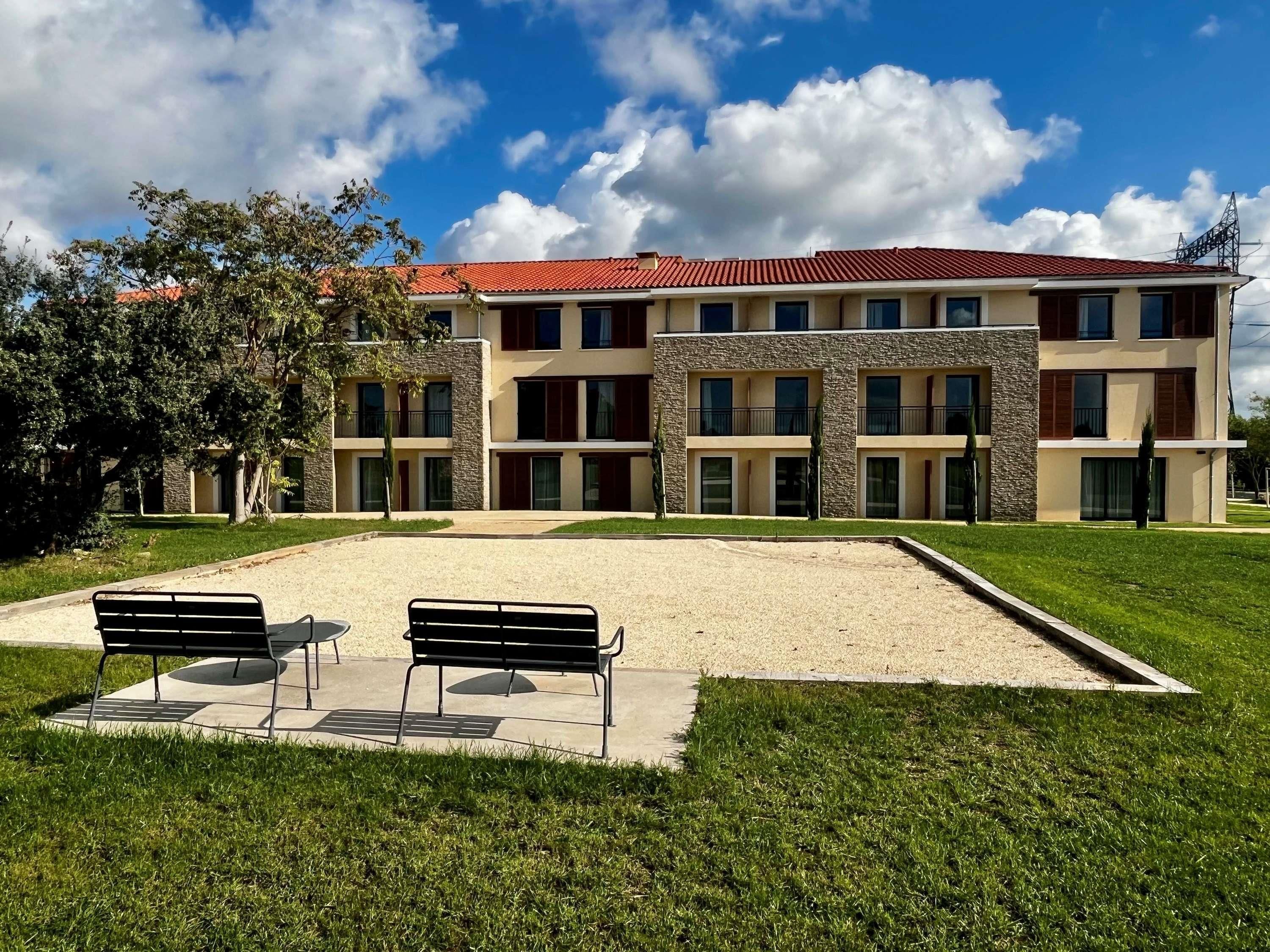 A multi-story building with a red-tiled roof, a courtyard with two benches, and a grassy area.