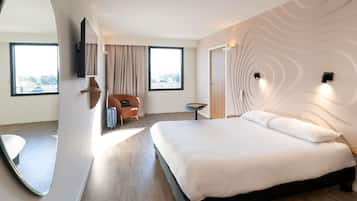 A modern hotel room with a large bed, a seating area with a chair and table, a large mirror, and a view of the city.