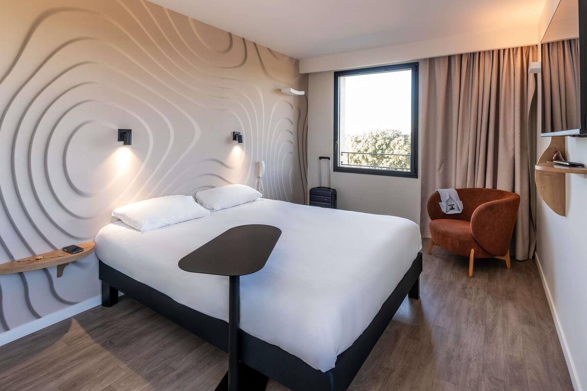 Une chambre d’hôtel moderne, équipée d’un lit, d’une petite table, d’une chaise, d’un téléviseur et d’une fenêtre avec des rideaux.
