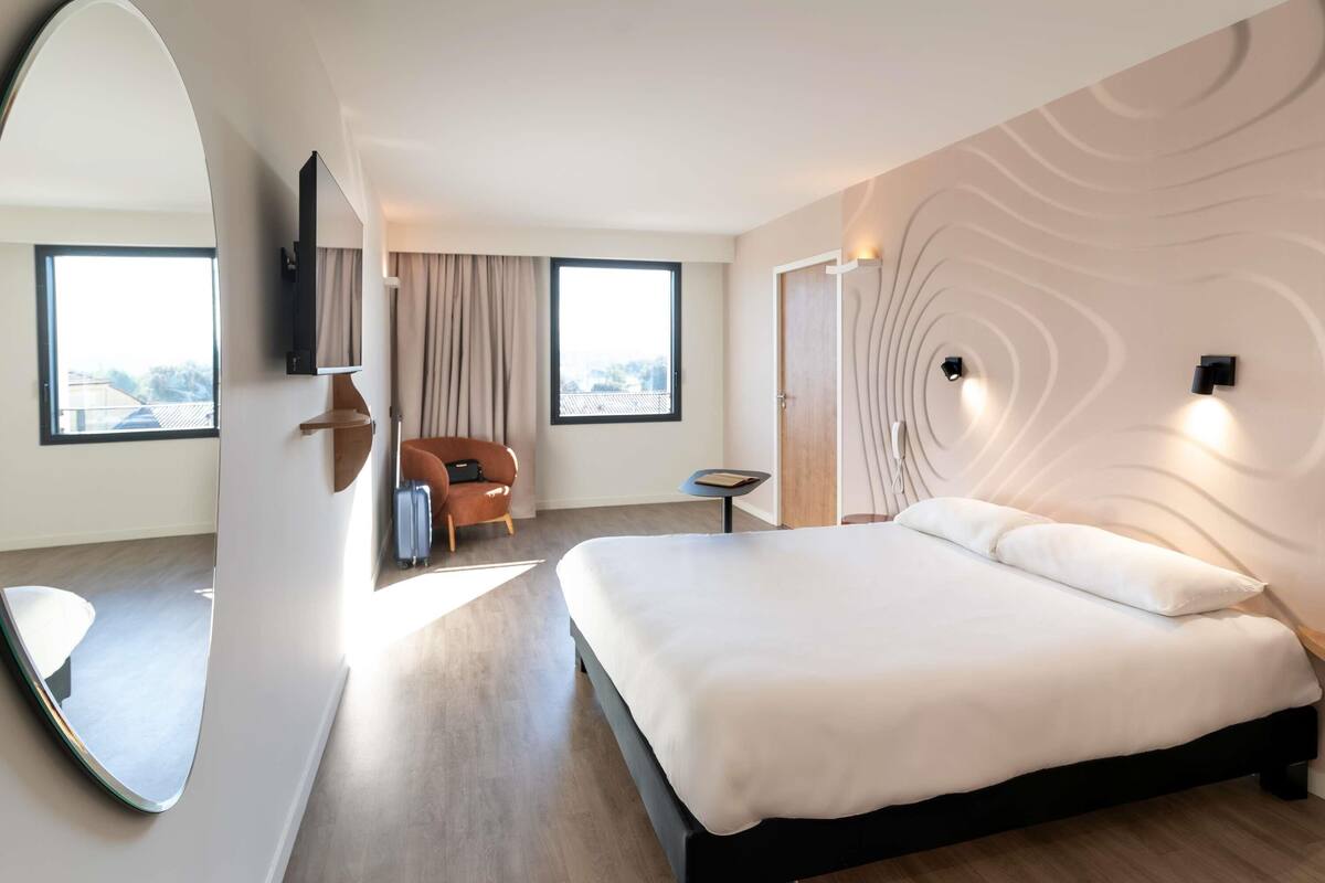 Une chambre d’hôtel moderne dotée d’un grand lit, d’un coin salon avec une chaise et une table, d ’un grand miroir et offrant une vue sur la ville.
