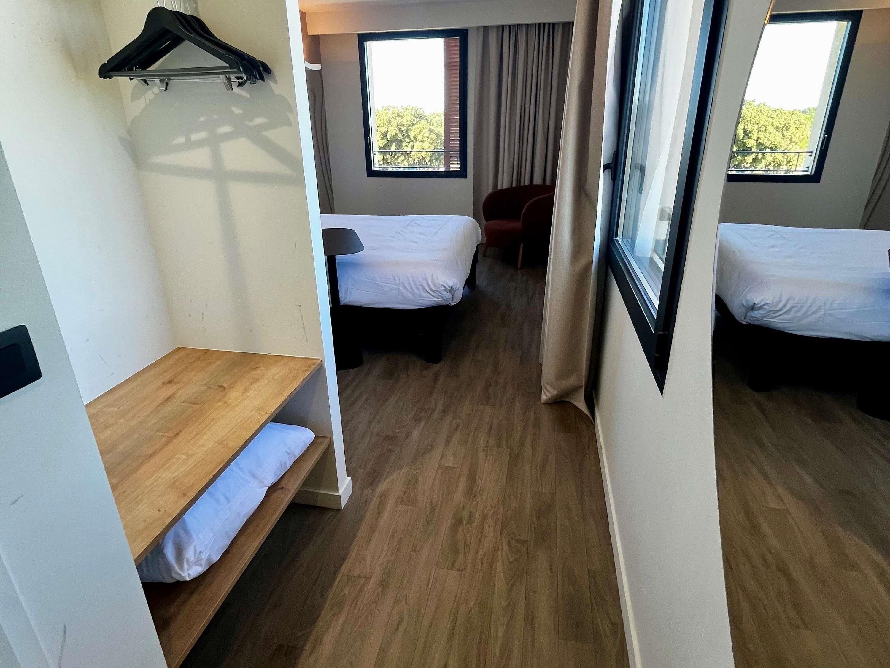 A hotel room with two beds, a wooden bench, a chair, and a window with a view of trees.