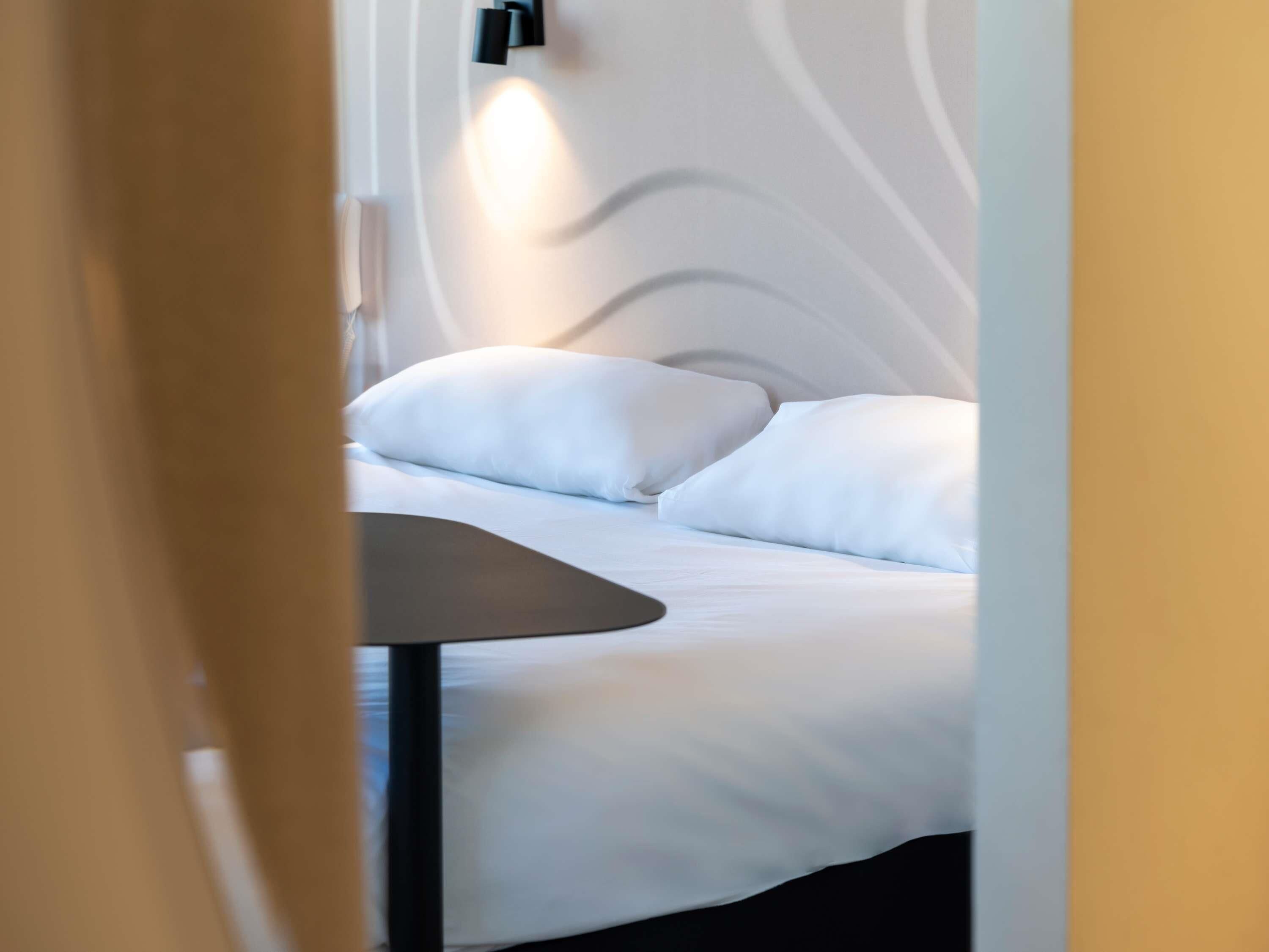 A modern hotel room with a neatly made bed, a small table, and a wall-mounted light fixture.