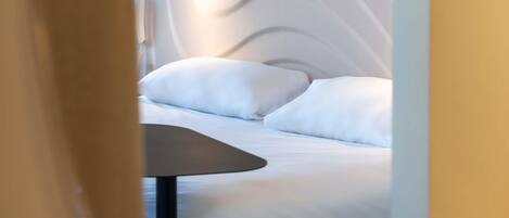 A modern hotel room with a neatly made bed, a small table, and a wall-mounted light fixture.