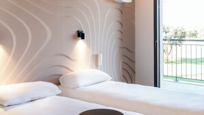 A hotel room with two beds, a bedside table, a phone, and a view of the outdoors. - Ibis Styles Cabriès Aix-En-Provence Tgv  (Cabries)