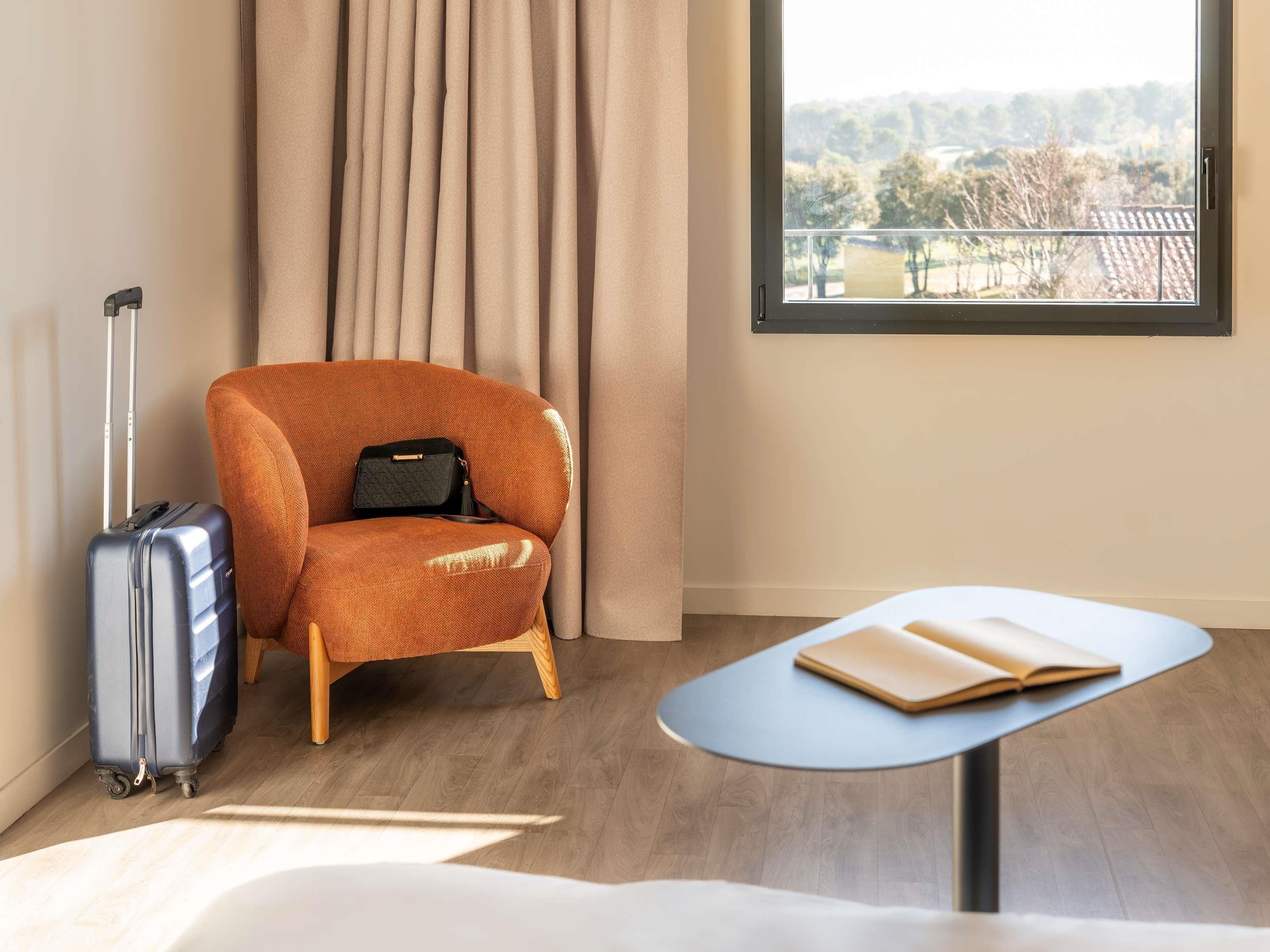 Une pièce avec un plancher en bois, une valise bleue, un fauteuil orange, une petite table avec un livre, et une fenêtre donnant sur des arbres et des immeubles.