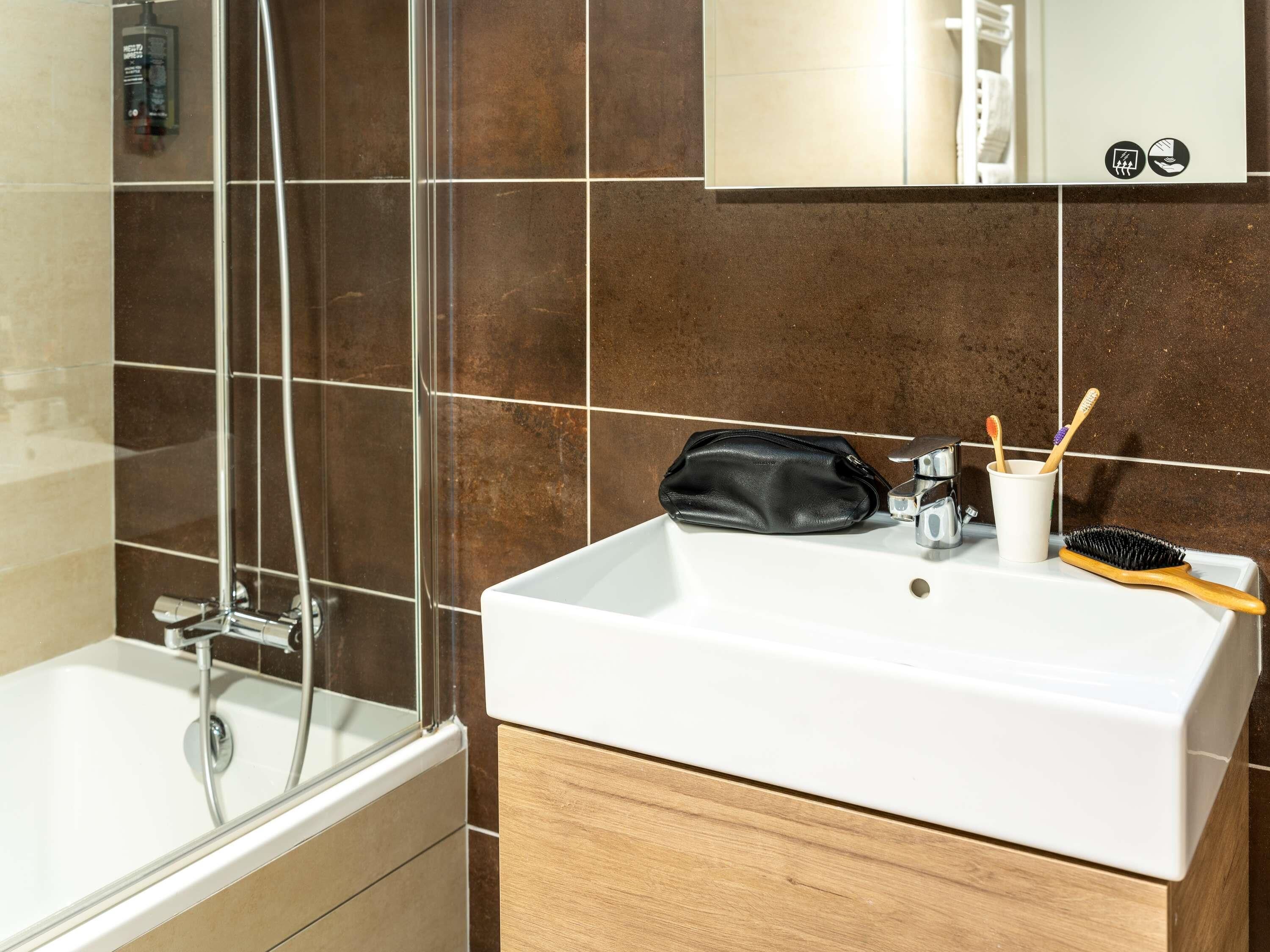 Un bagno con un lavandino bianco, una borsa nera, una tazza con gli spazzolini e uno spazio doccia.
