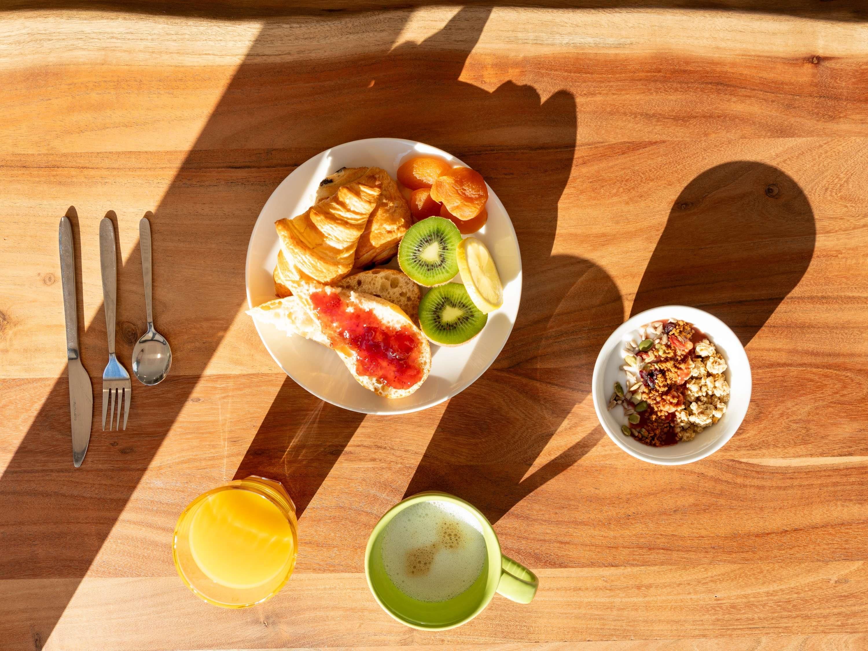 Une table en bois dressée pour le petit-déjeuner, avec des croissants, des tranches de kiwi, de la confiture, un bol de granola, un verre de jus d’orange et une tasse de café.