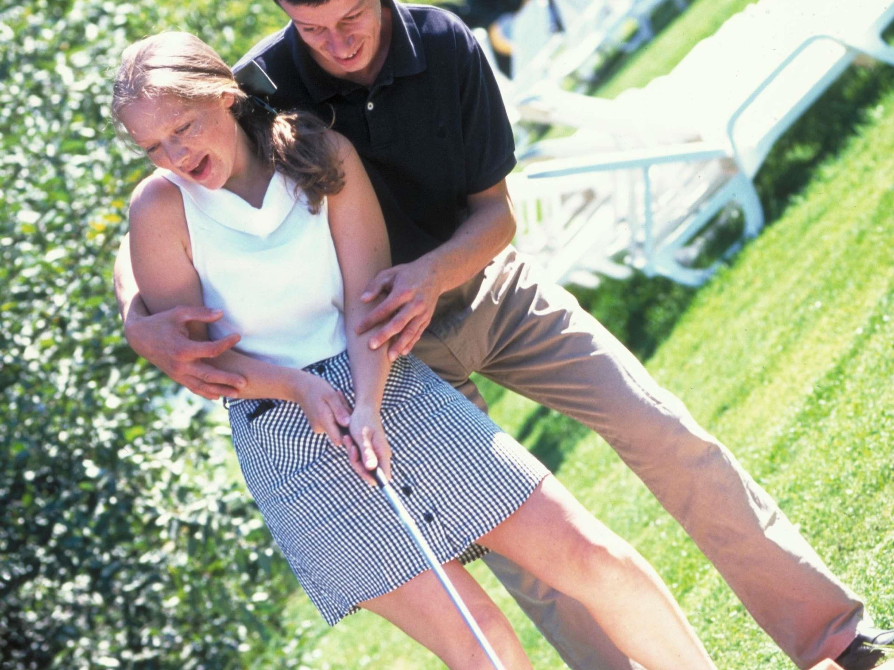A person in a white sleeveless top and checkered skirt standing on a grassy area with a golf club.