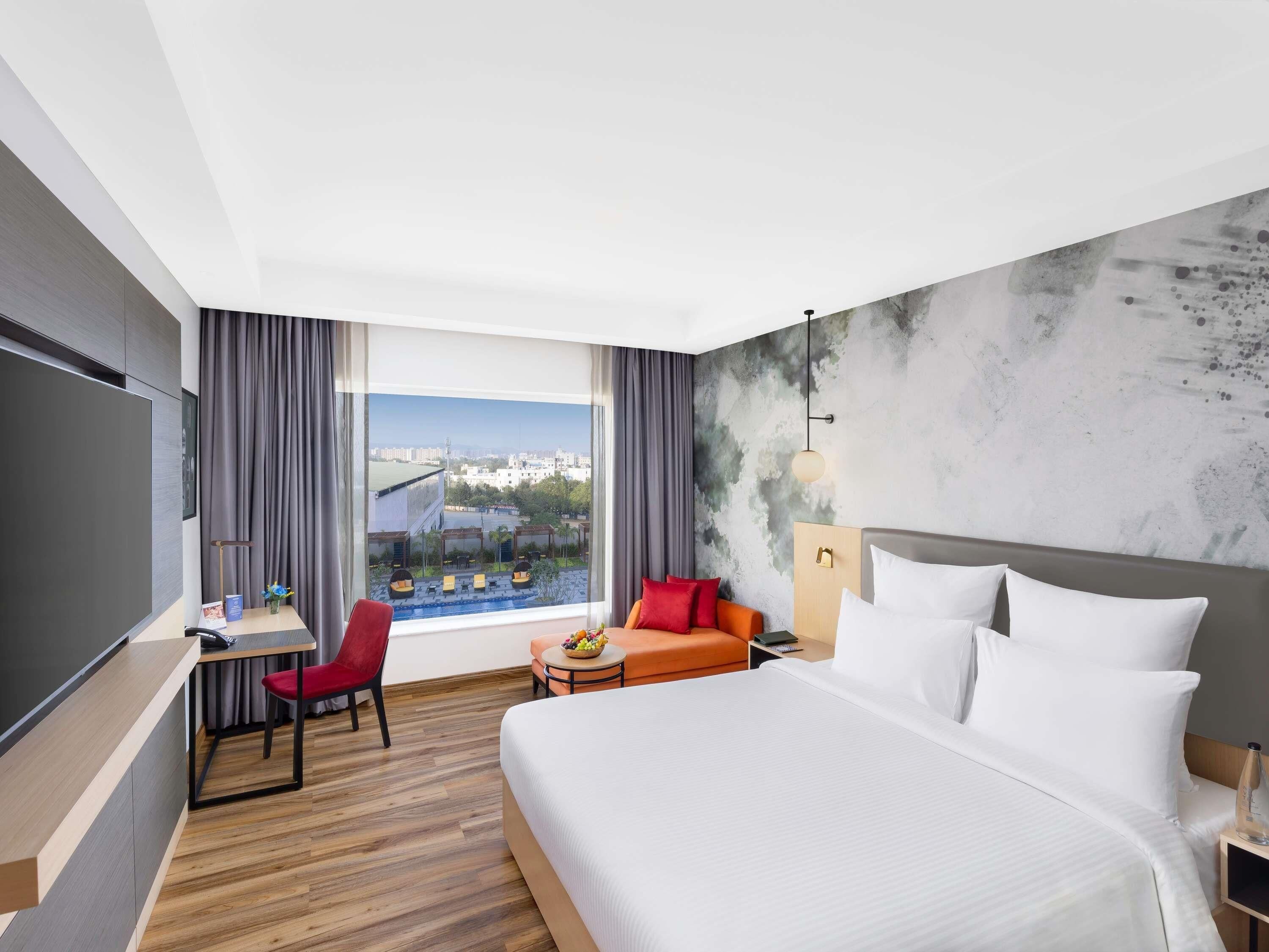A hotel room with a bed, a desk, a chair, a small table, and a view of a pool and buildings.
