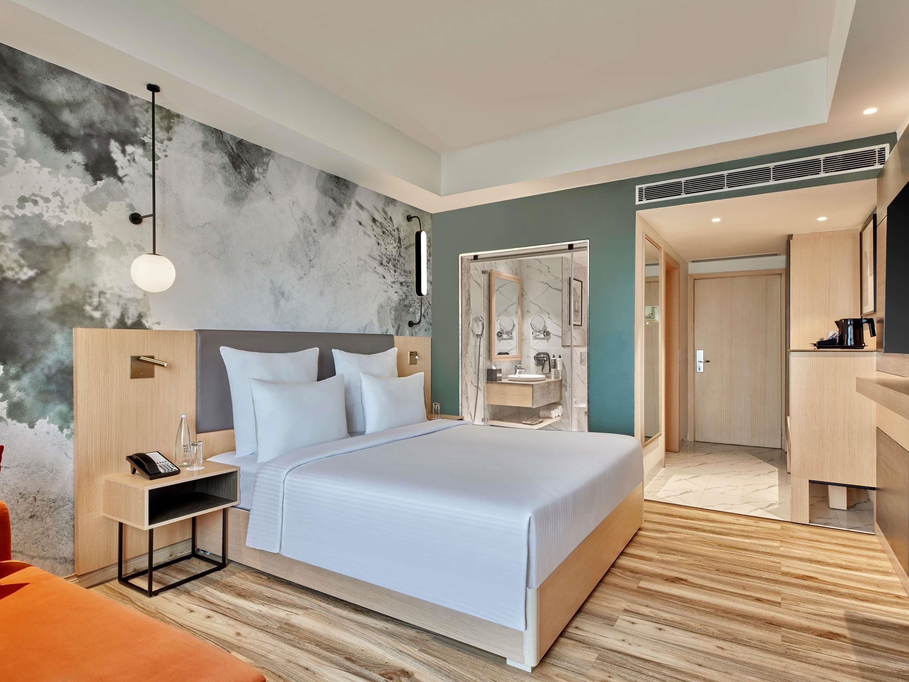 Una habitación de hotel moderna con una cama grande, una mesita de noche con teléfono, un espejo y un baño con ducha e inodoro a la vista.