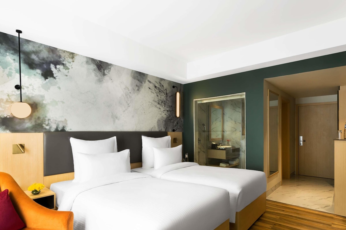 A hotel room with two beds, a nightstand with a lamp, a wooden floor, and a bathroom visible through a glass door.
