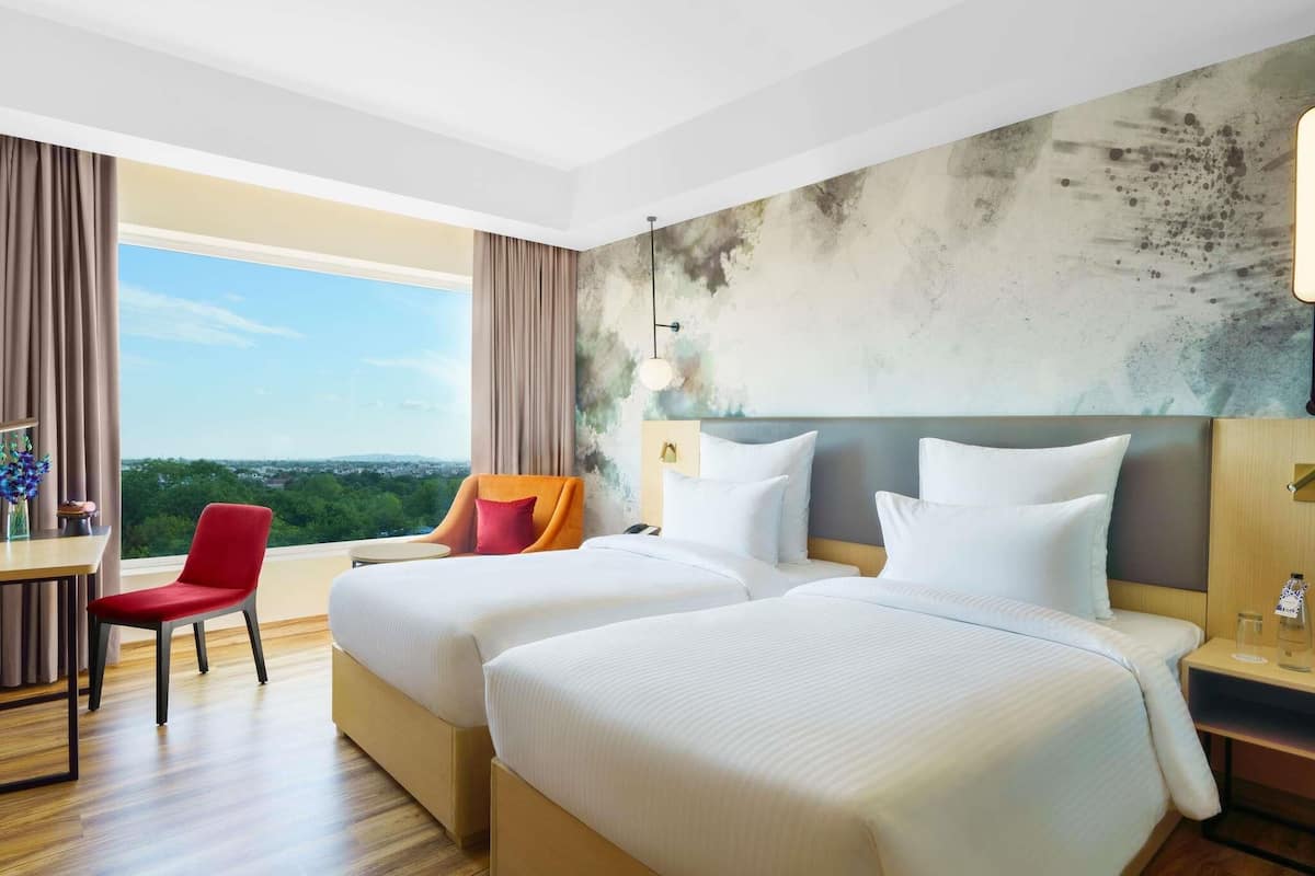A hotel room with two beds, a desk, a chair, and a large window with a city view.