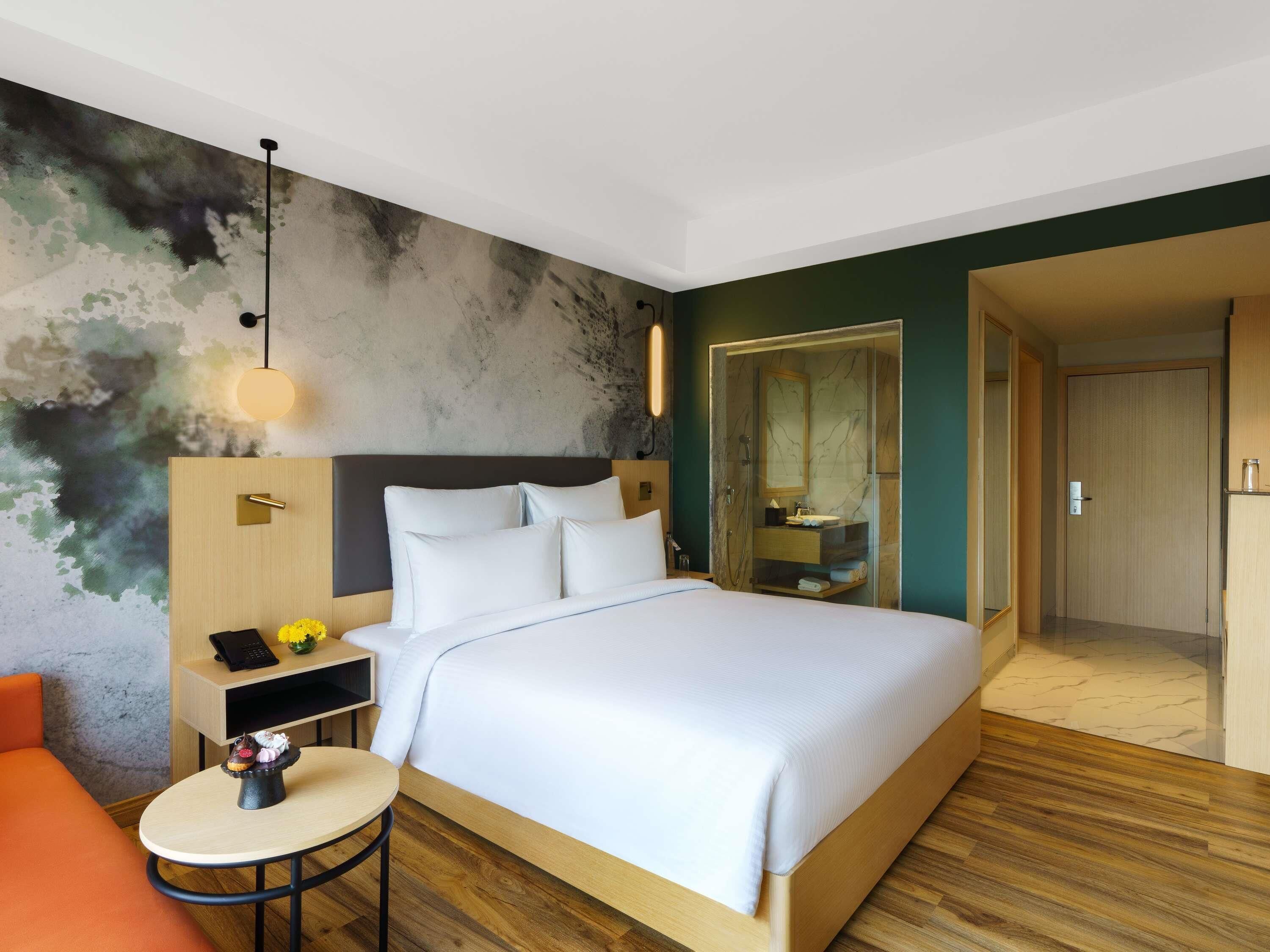 A modern hotel room with a large bed, a bedside table with a lamp, a small round table with a vase of flowers, and a view of the bathroom through a glass door.