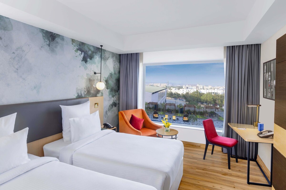 Habitación de hotel con dos camas, un escritorio, una silla, vista a la ciudad y un diseño de techo moderno.