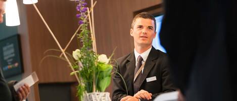 A man in a suit seated at a table with a name tag, a vase with flowers, and a modern interior with pendant lighting.