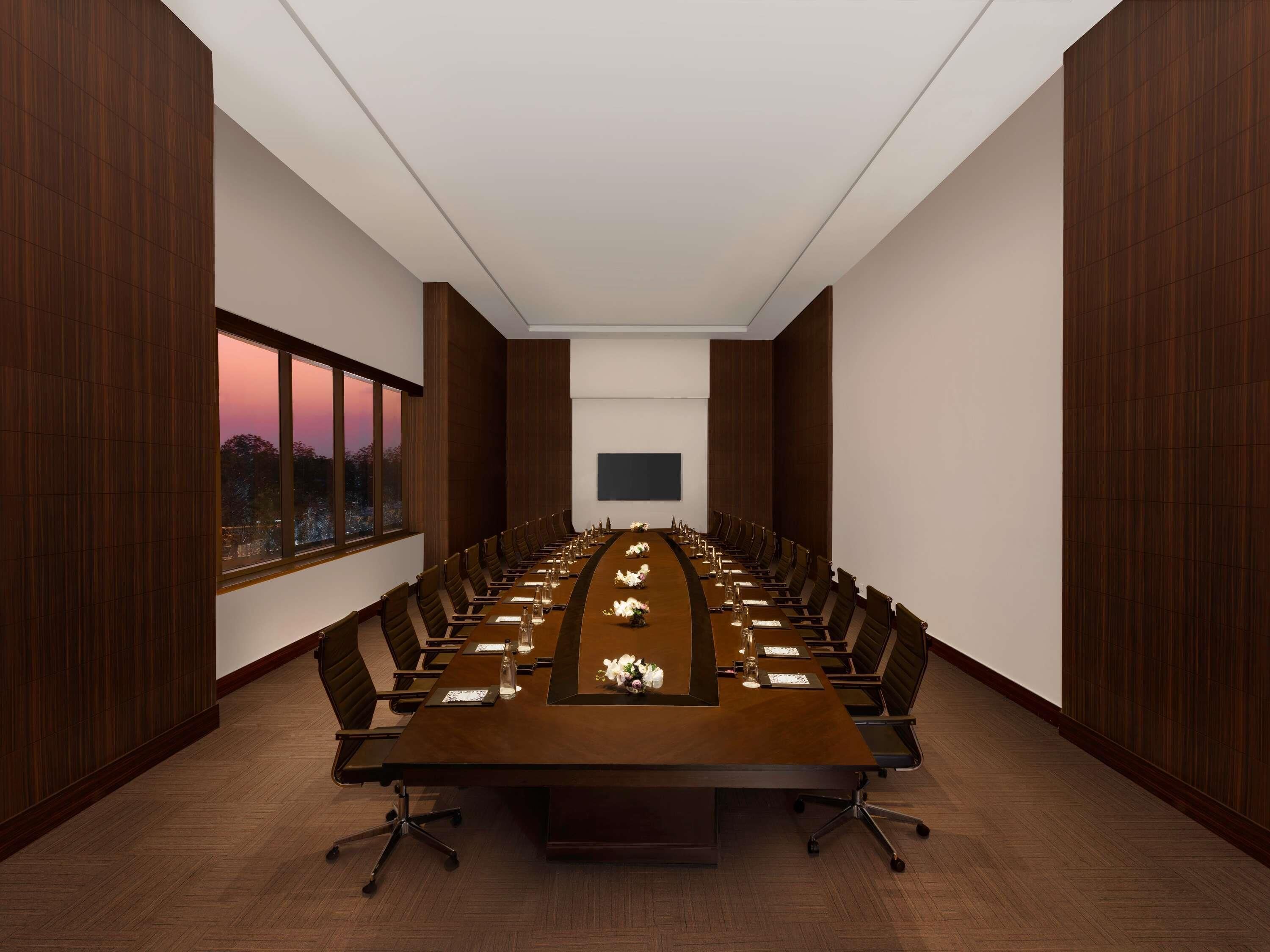 A conference room with a long table, chairs, a projector screen, and a view of the sunset through large windows.