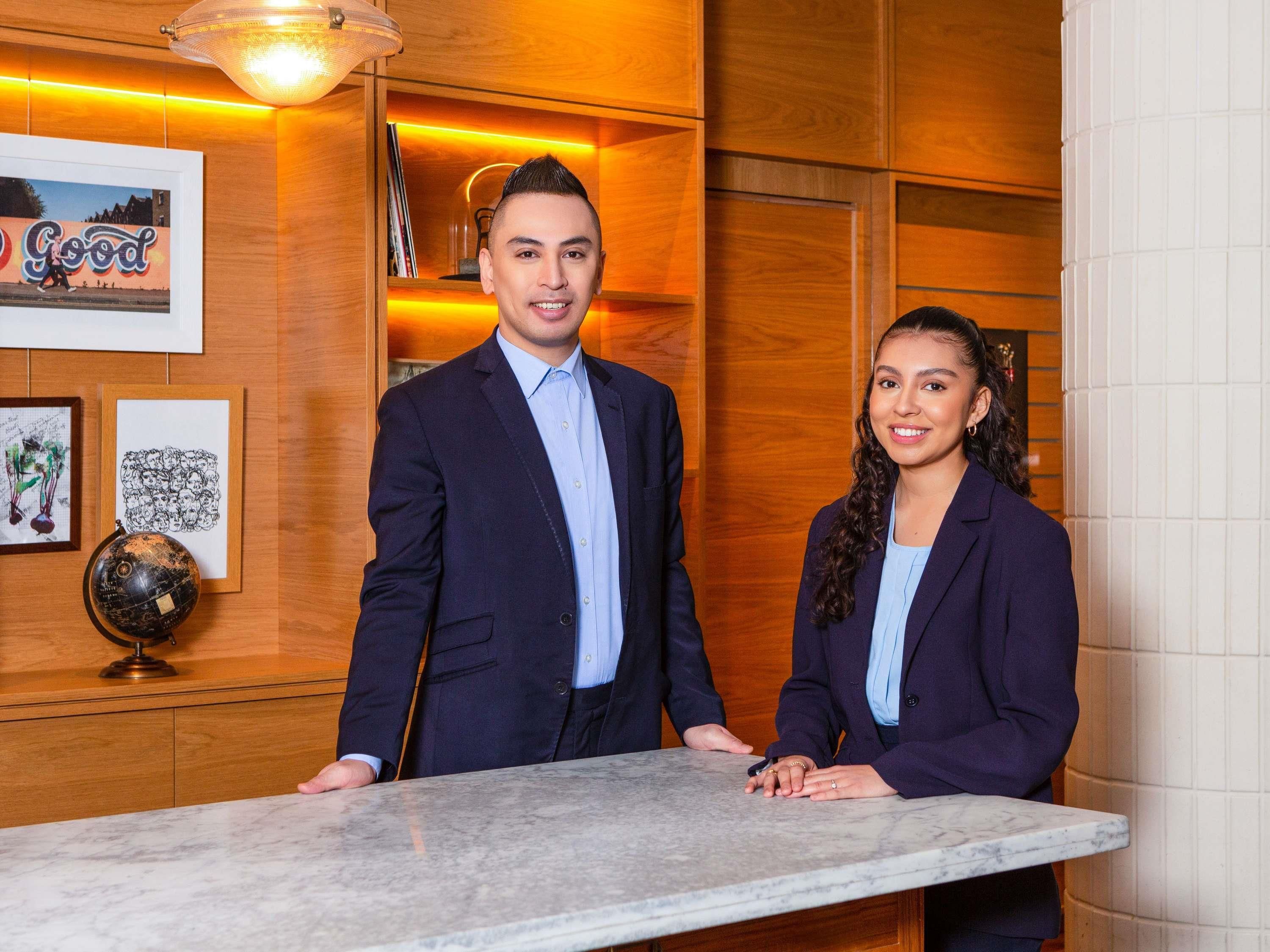Two individuals standing at a reception desk in a hotel lobby with wooden paneling and framed artwork on the walls.