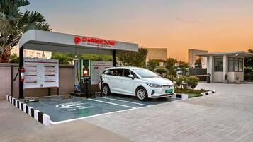 An electric vehicle charging station with a white car parked, a sign with instructions, and a building in the background.