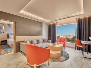 Une chambre d’hôtel moderne, dotée d’un canapé, de fauteuils, d’une table à manger et d’une grande fenêtre donnant sur la ville.
