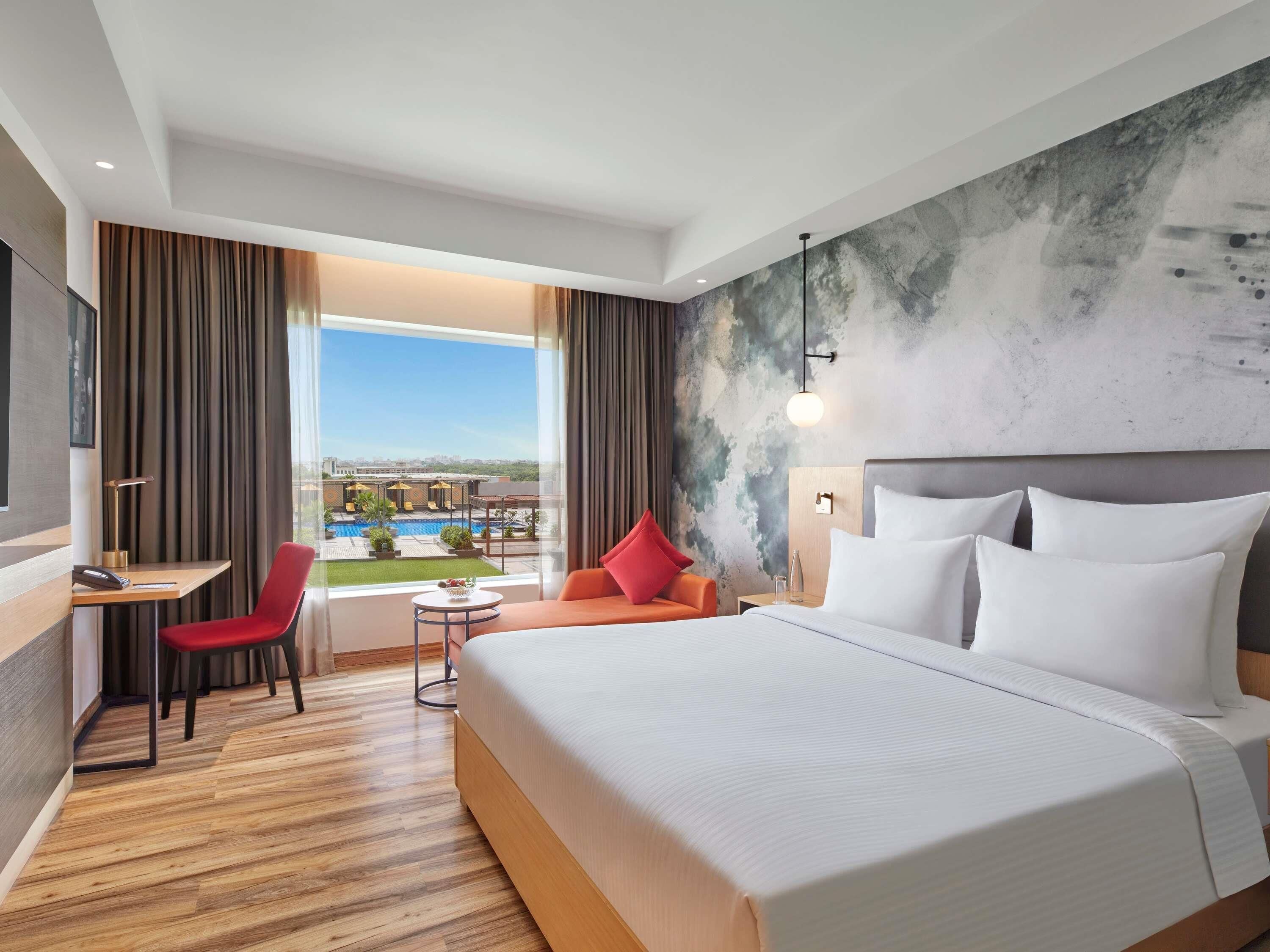 Een hotelkamer met een groot bed, een bureau, een stoel en uitzicht op een zwembad en gebouwen.