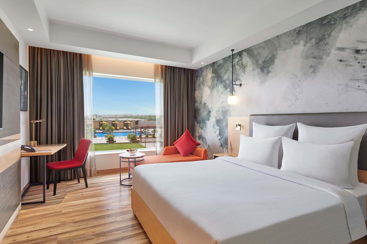 A hotel room with a large bed, a desk, a chair, and a view of a pool and buildings.