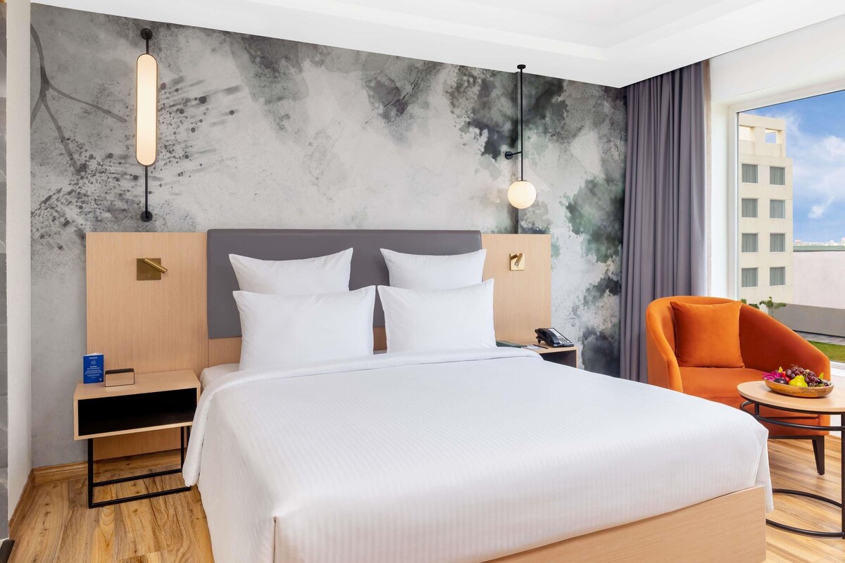 Habitación de hotel con una cama grande, una mesita de noche de madera, un escritorio, una silla y vistas a edificios a través de la ventana.