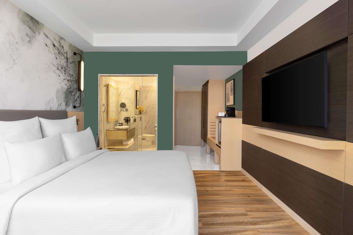 A modern hotel room with a large bed, a TV mounted on the wall, a bathroom with a glass shower enclosure, and a wooden floor.