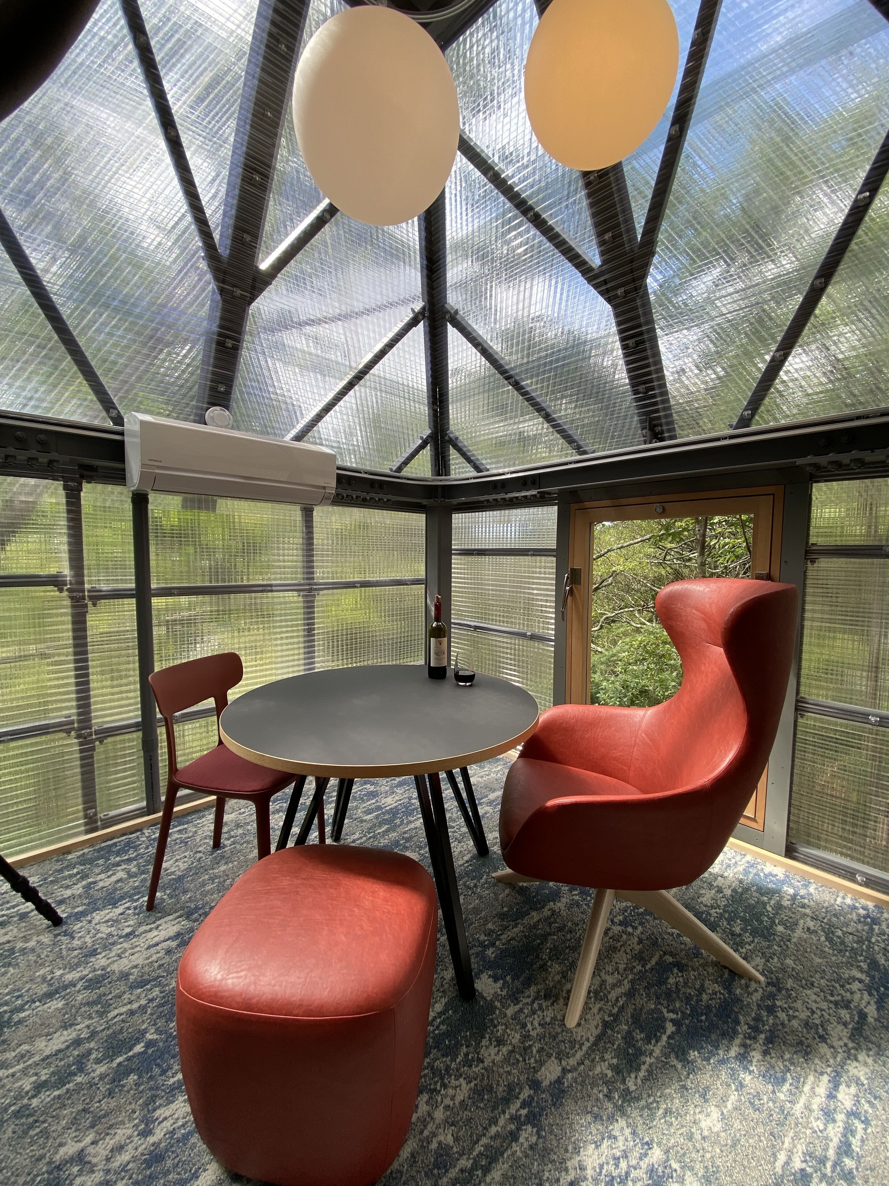 Une pièce avec une table ronde, deux chaises et un pouf rouge. Elle est dotée d’une grande verrière et offre une vue sur la verdure environnante.