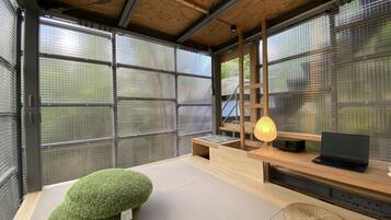 A modern room with large windows, a laptop on a wooden desk, a green cushion, and a woven mat.