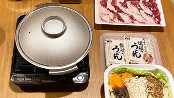 A portable electric stove with a lid, a container of soy sauce, a plate of sliced meat, and a bento box with vegetables and mushrooms.