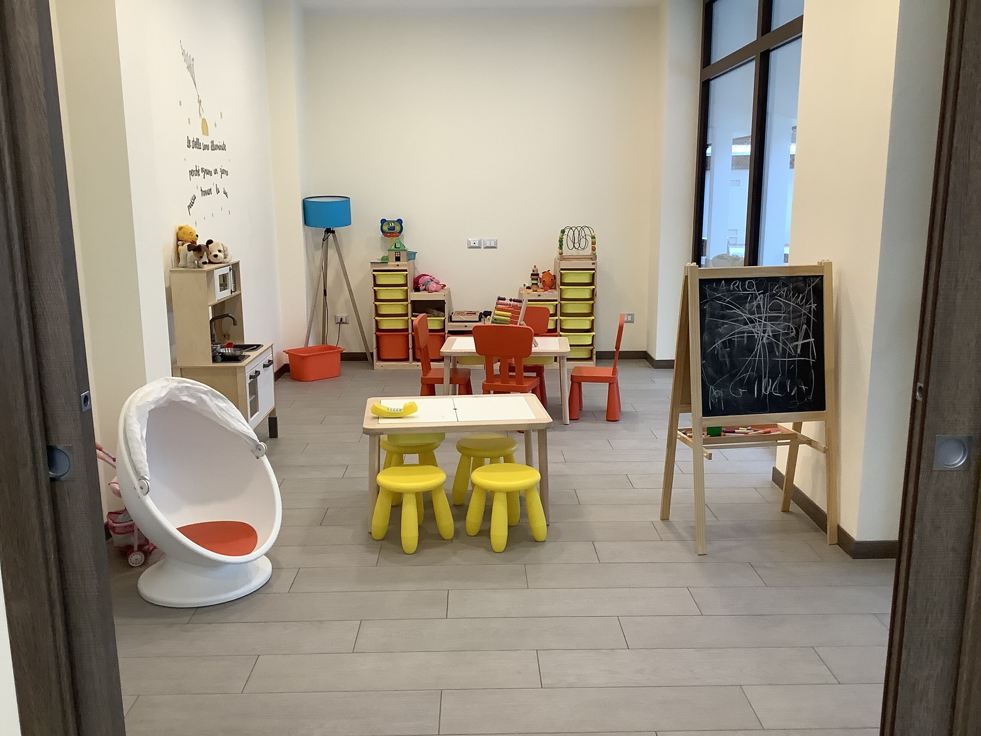 A children's playroom with a white chair, yellow stools, a chalkboard, and storage units.