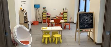 Una sala giochi per bambini con una sedia bianca, sgabelli gialli, una lavagna e contenitori portaoggetti.