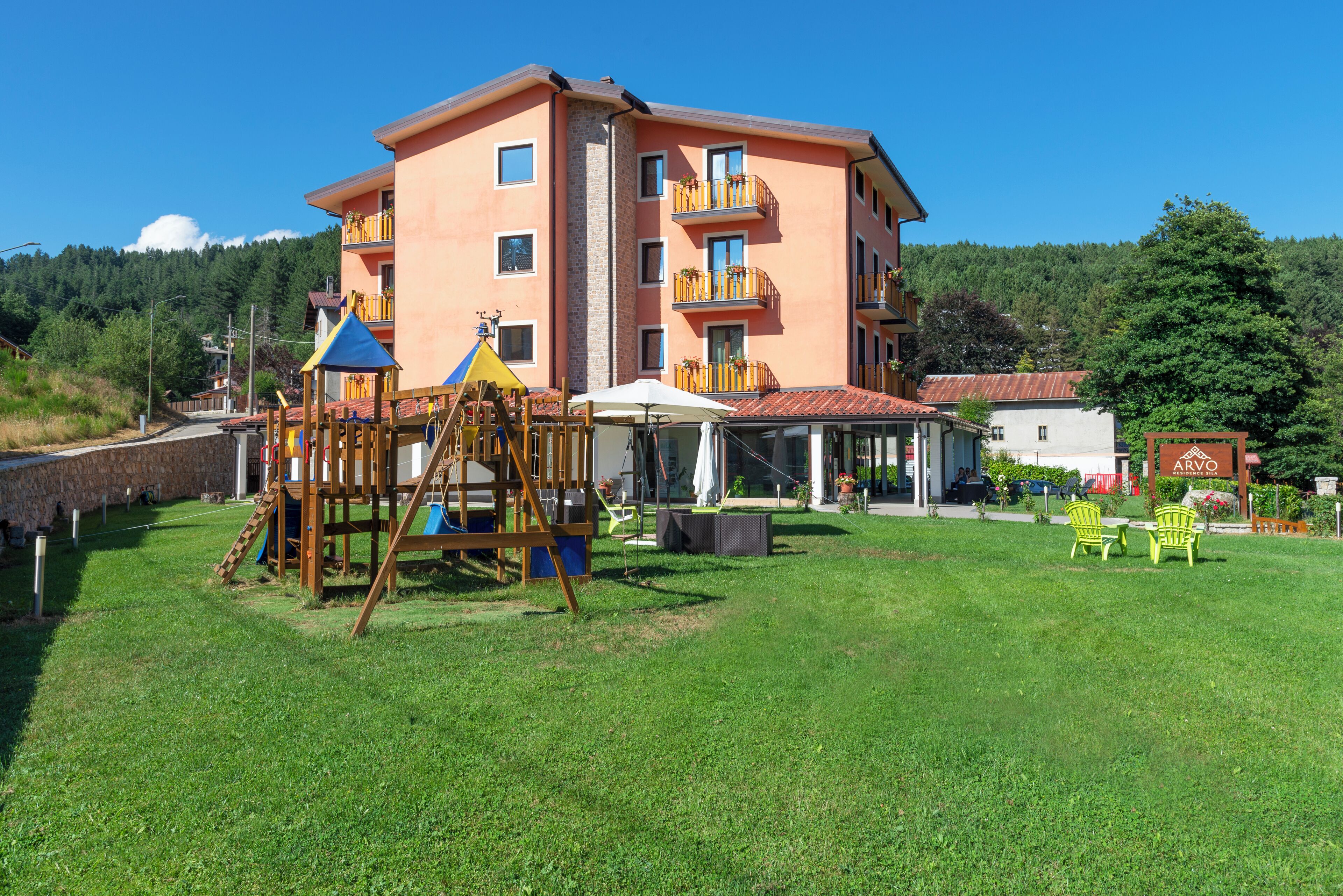 Un edificio de varios pisos con un parque infantil en primer plano, una zona de estar al aire libre cubierta y un cartel que dice 'ARVO'.