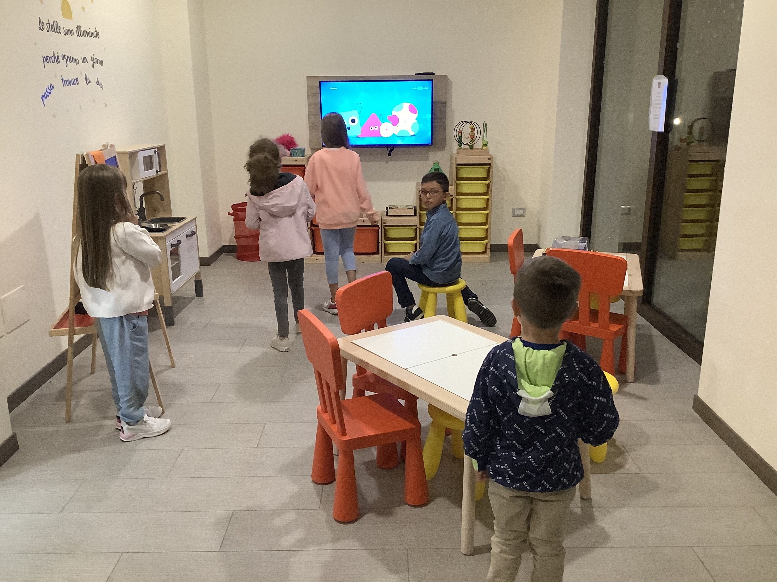 Un groupe d’enfants et d’adultes se trouve dans une pièce où l’on peut voir un téléviseur diffusant des formes colorées, une table avec des chaises et des meubles de rangement dotés de bacs jaunes.