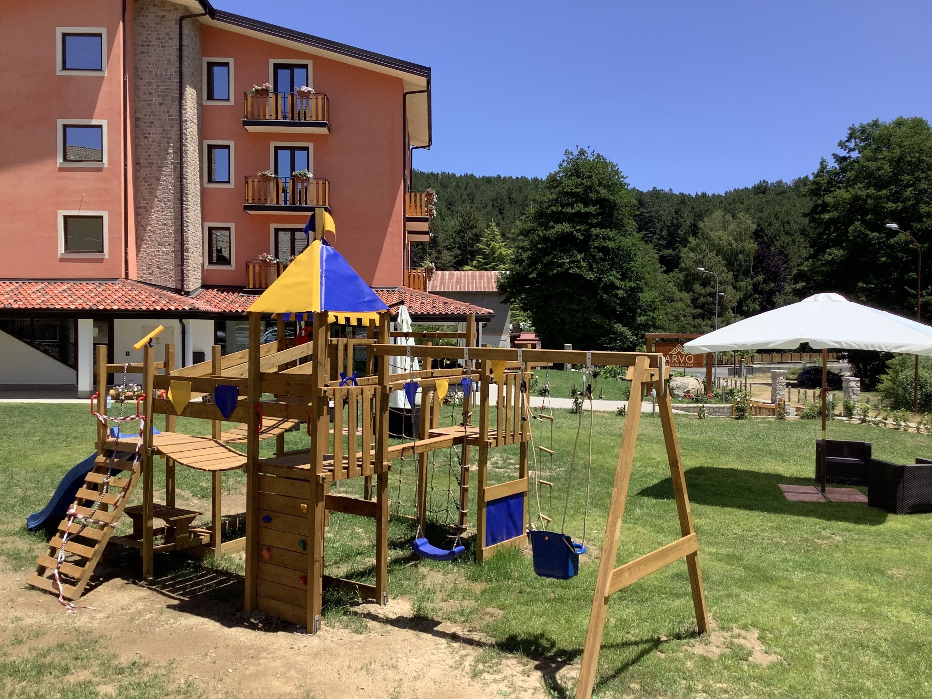 A playground with a wooden structure, including a slide and swings, in front of a building with multiple windows and balconies.