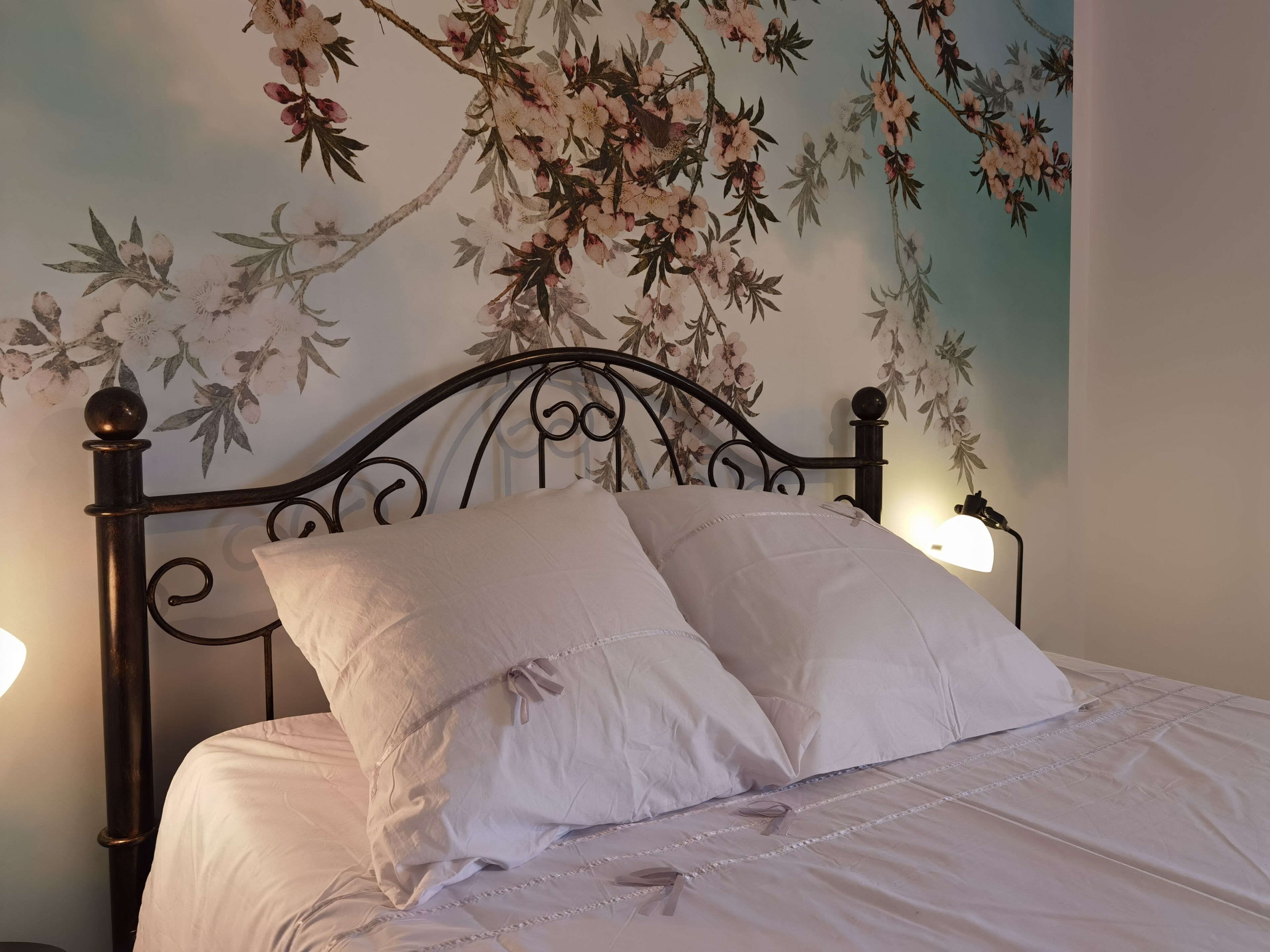 Uma cama bem arrumada com lençóis brancos e duas almofadas, cabeceira de metal escura e uma parede com um mural floral.