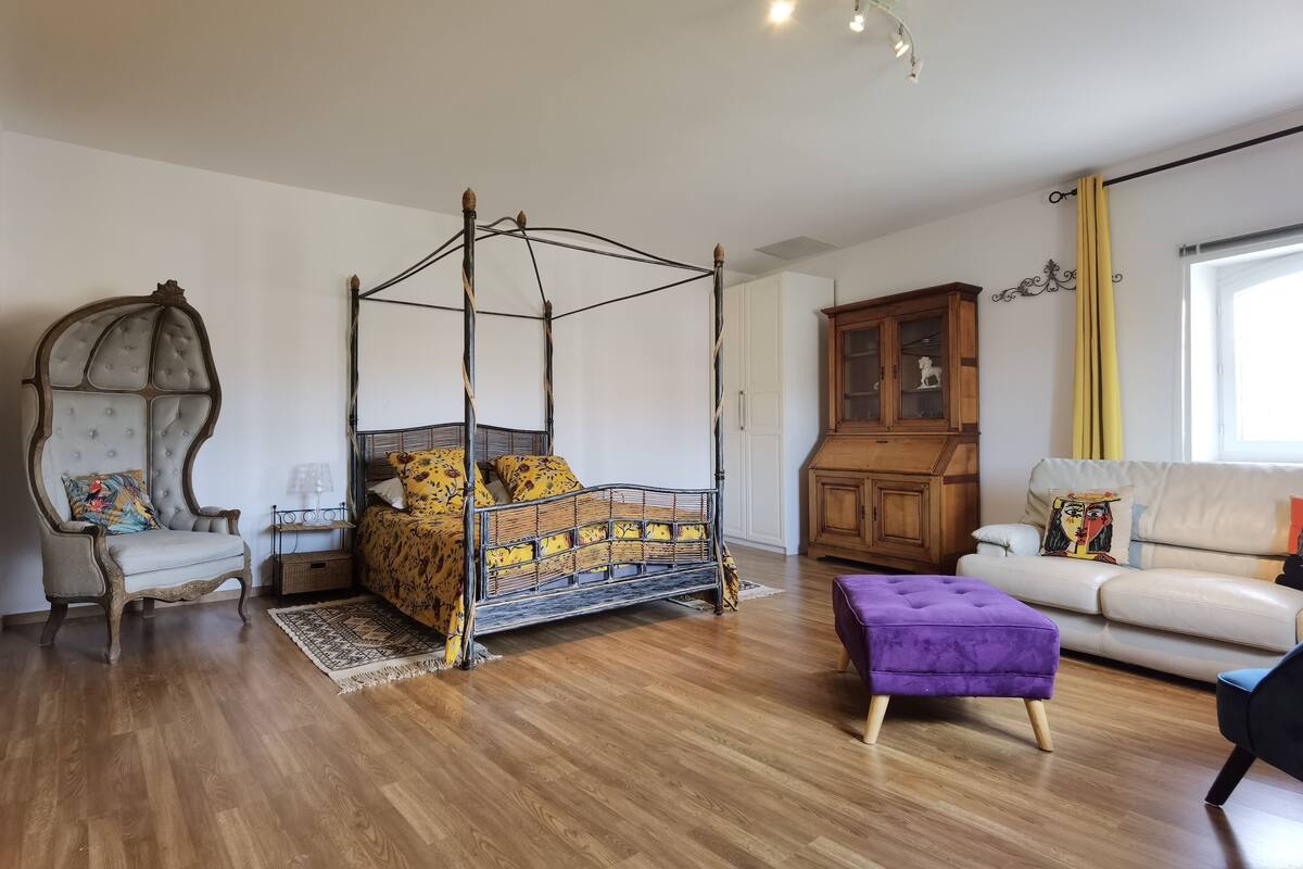 Ein geräumiger Raum mit einem Himmelbett, einem Holzschrank, einem Sofa und einem violetten Ottomanen.