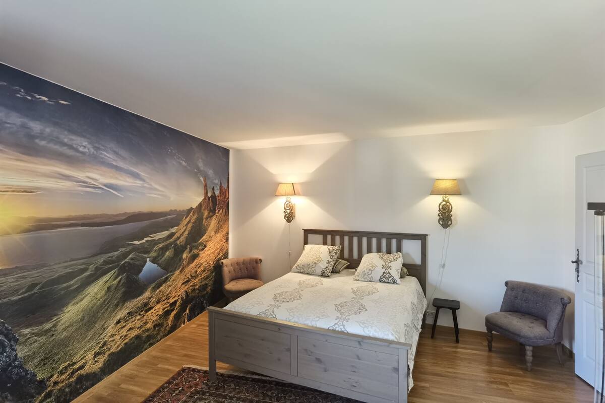 Ein Schlafzimmer mit einem großen Landschaftsbild, einem Holzbett, zwei Wandleuchten und einem Sessel.